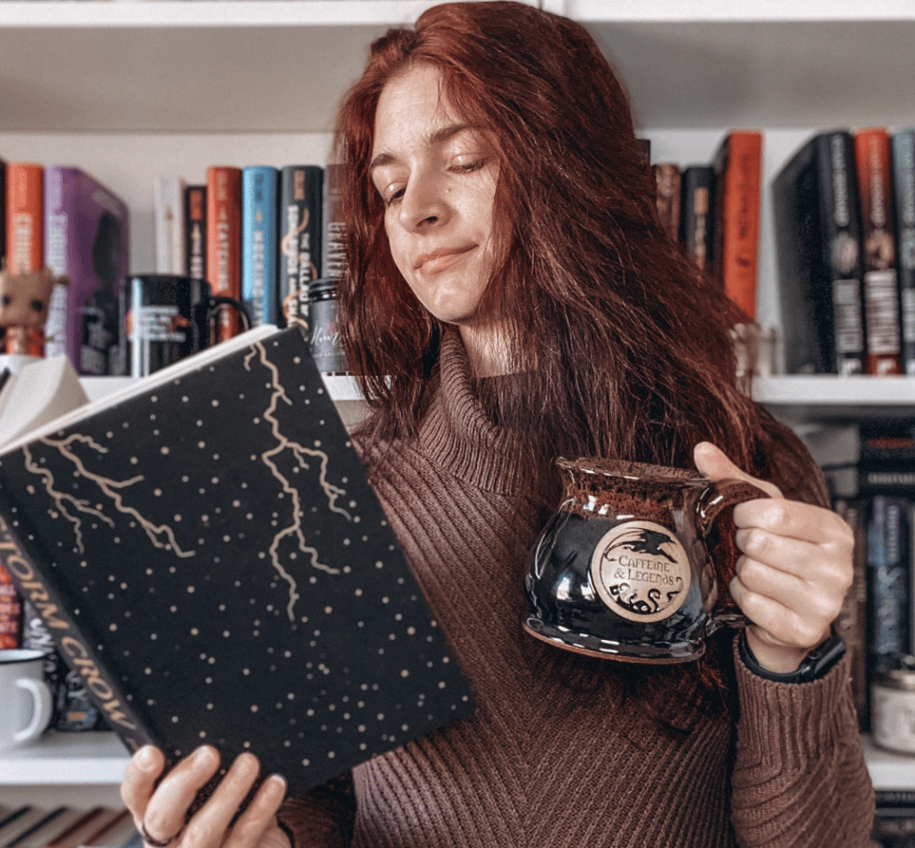 A young woman standing in front of her bookshelf, holding a book and a mug.