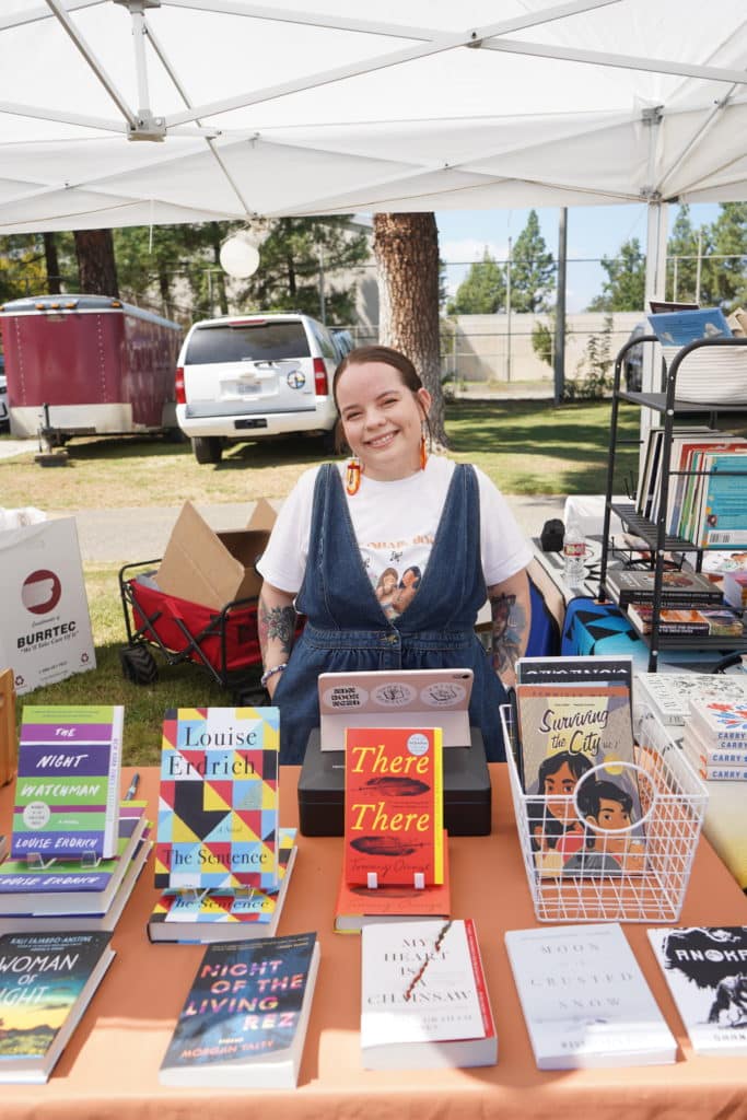 A young woman is standing behind a table of books, and smiling.