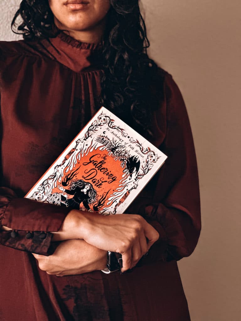 A woman with curly hair is standing close to the camera, and is cradling a book against her chest.