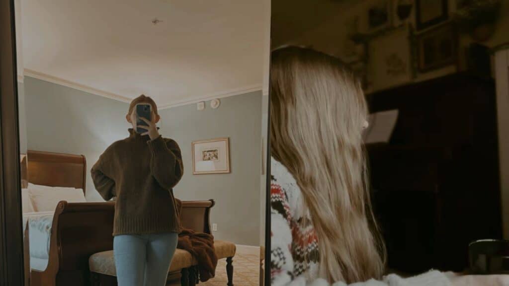 Two images side by side.On the left, our Bookstagrammer of the Week, Madison, is taking a mirror selfie. Her phone obscures her face but we can see that she is wearing a dark brown sweater with light gray bottoms. Her bed is visible in the background and it is dressed with white covers and pillows. There is an ottomon at the foot of her bed with a plush looking bunch of fabric in the middle of it. On the right, we can see the side profile of Madison. She is completely obscured from view by her long blonde hair. She is wearing a brightly patterned cardigan. cr. Madison / @classic.literature.love
