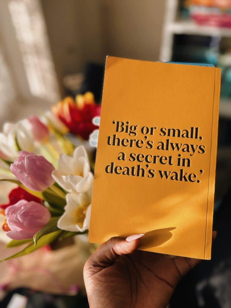 Our Bookstagrammer of the Week, Anita, holds a yellow book against a bouquet of flowers. The flowers are pink, white, red and yellow tulips. The book has a large quote on it in a serif font. The quote reads: "Big or small, there's always a secret in death's wake." This is the back cover of The Things That We Lost by Jyoti Patel.Image credit to Anita of @booksinherhands.