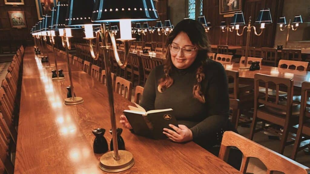 Our Bookstagrammer of the Week, Matina of @breathing_pages, is sitting in a library reading a hardcover book by lamplight. She is wearing a dark long sleeved shirt and thin-rimmed glasses. cr. Matina / @breathing_pages