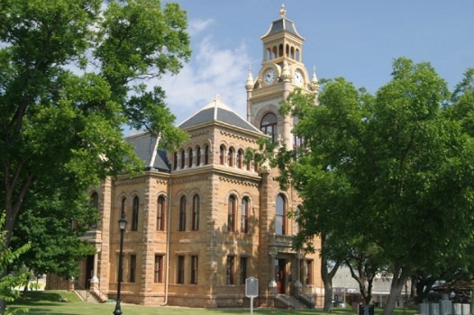 A large brick clock tower and building sit behind two trees. This is a photo of Llano County's Courthouse.Image credit Llano County Texas website
