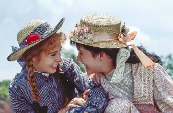 Anne Shirley and Diana Barry laughing while looking at each other, they are both wearing dresses and hats with a flower decoration - Anne of Green Gables series (year 1972)