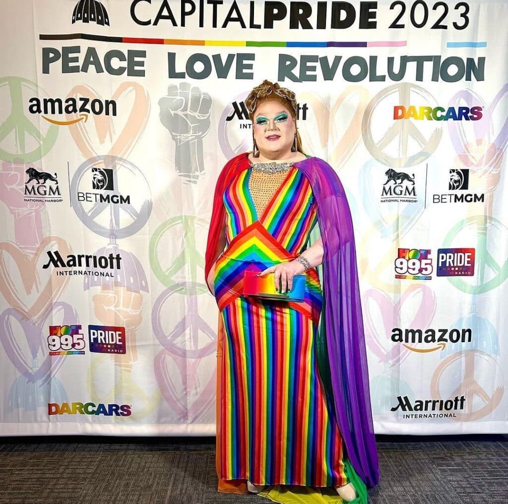Drag queen wearing rainbow dress, standing against background that says "Capital Pride 2023" and under that, "Peace Love Revolution"