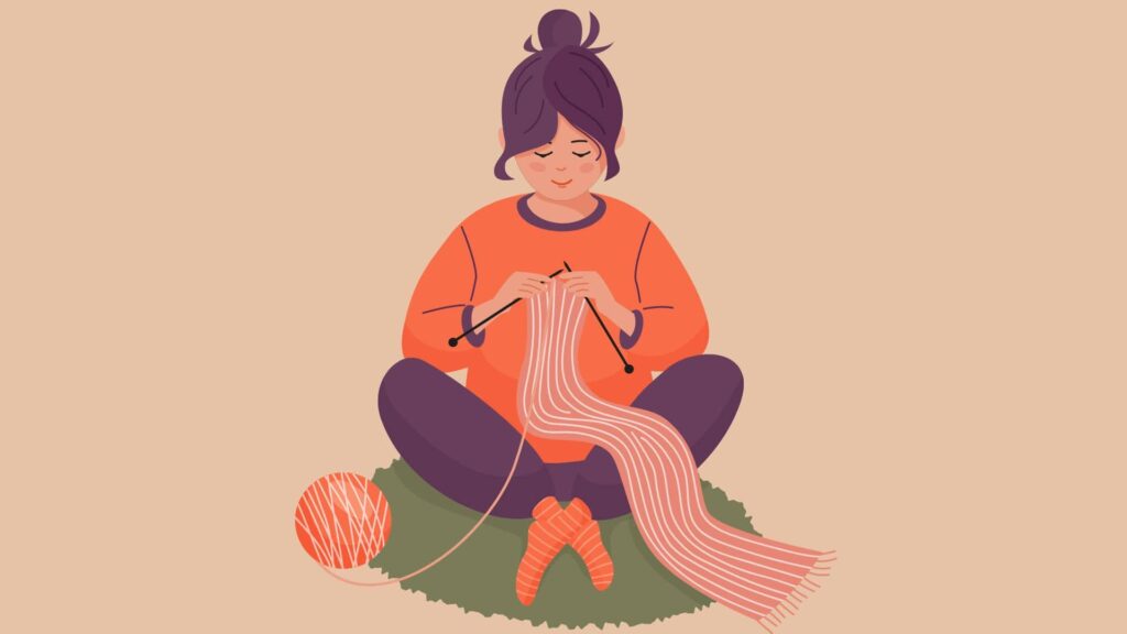 background-is-tan-and-in-front-of-it-is-a-woman-sitting-cross-legged-in-orange-long-shirt-and-black-pants-knitting-a-lighter-orange-scarf