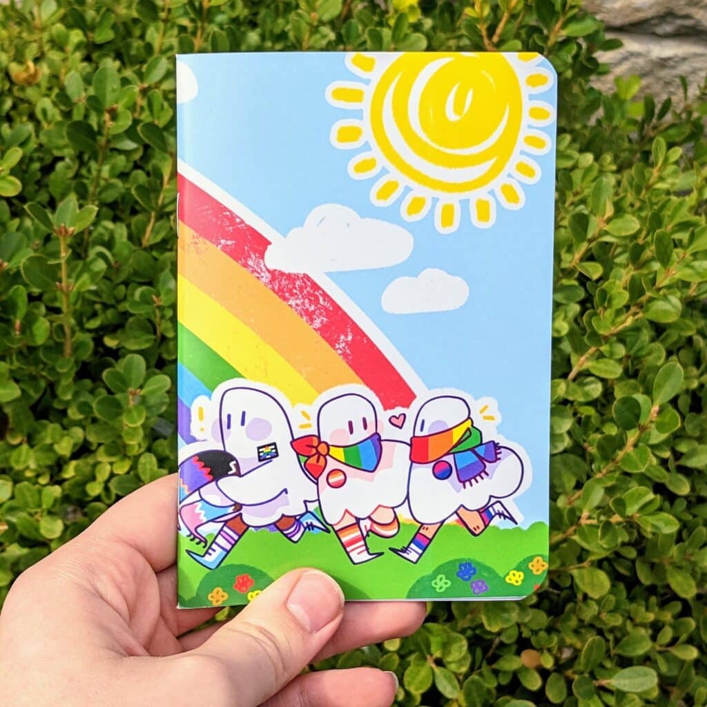 5-inexpensive-gifts-to-surprise-your-favorite-queer-writer-small-notebook-with-ghosts-wearing-lgbtqia-pride-clothes-on-sketchy-natural-background-being-held-over-a-bush