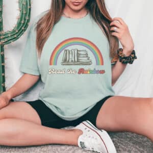 10-tempting-gifts-queer-book-nerds-will-love-girl-sitting-wearing-green-shirt-with-read-the-rainbow-and-a-rainbow-over-a-stack-of-books-on-it