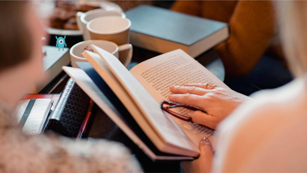 books-cups-and-food-on-a-round-black-table-and-there-are-women-reading-books-around-it. there-is-a-blue-tbr-monster-on-a-book