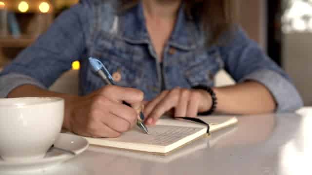 closeup-image-of-a-woman-writing-in-a-journal-on-a-table-next-to-a-coffee-cup