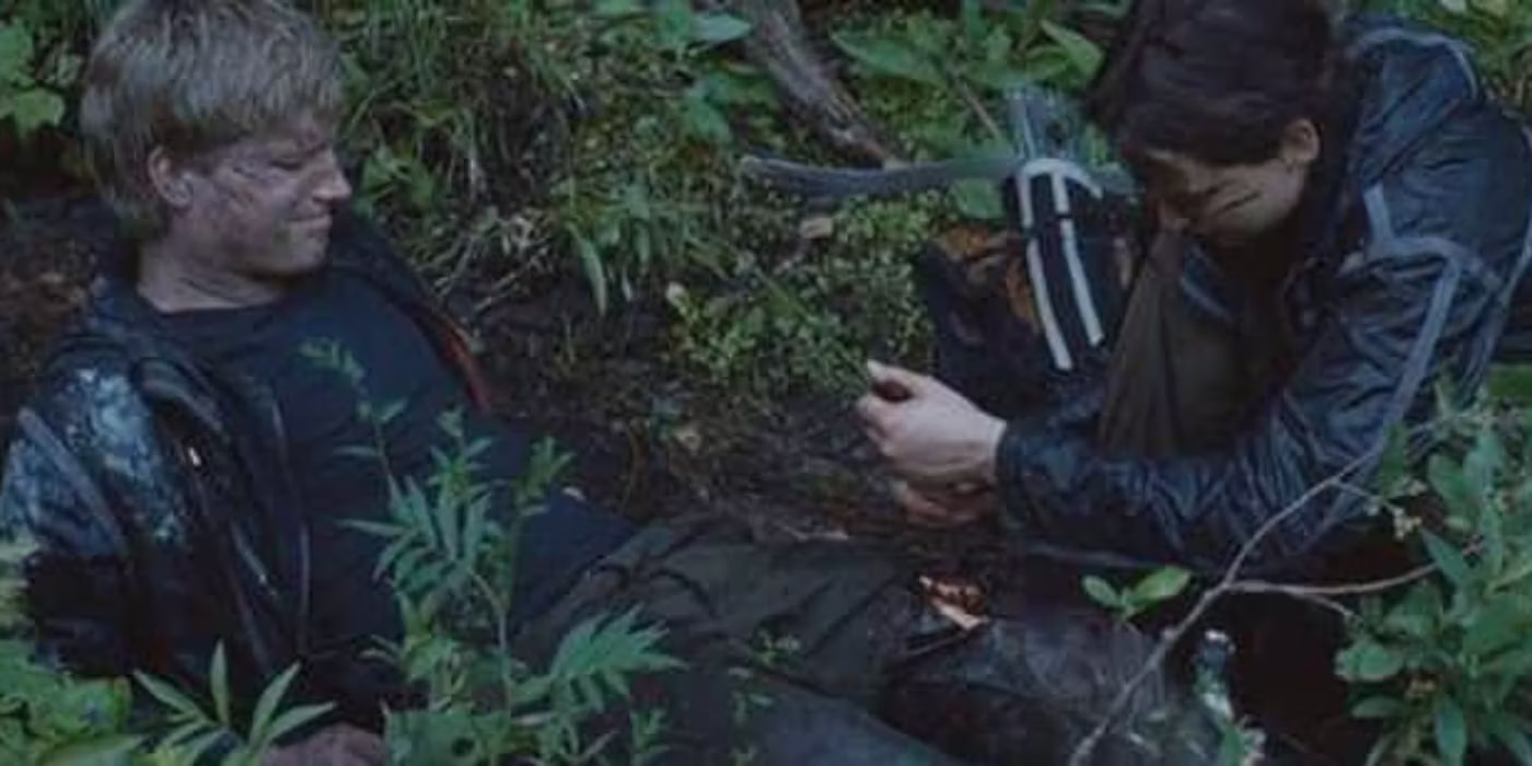 Peeta is lying on the forest floor in pain while Katniss tends to his injured leg
